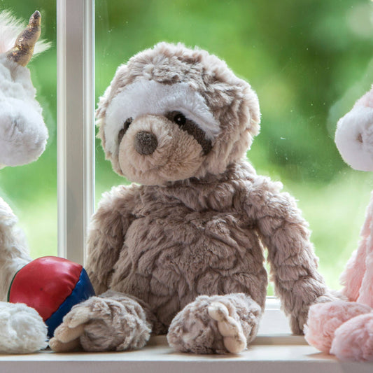 The Mary Meyer Putty Nursery Sloth, a machine washable plush with gray and white fur, sits on a windowsill holding a red and blue ball—blurred stuffed animals flank it, greenery outside—an ideal charming gift for babies.