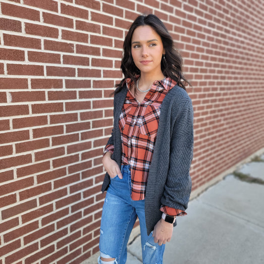 A young woman with long dark hair stands by a brick wall wearing the Staccato Macie Cardigan over a red plaid shirt and ripped blue jeans, looking to the side with a neutral expression.