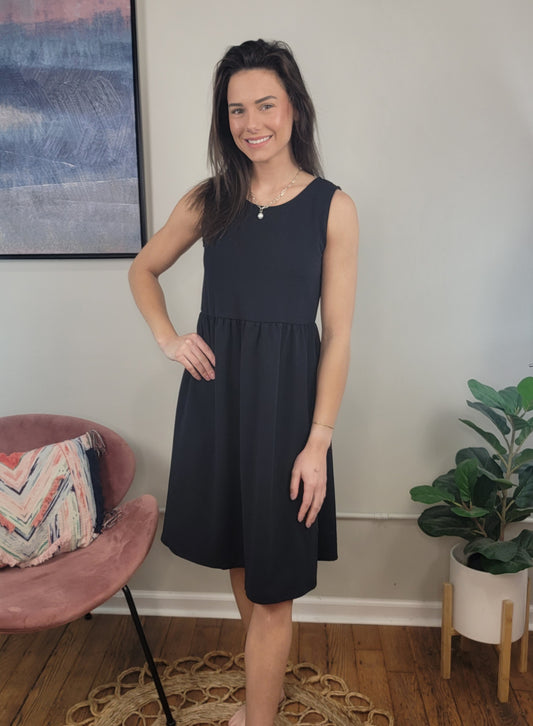 A woman with long brown hair smiles indoors, wearing the sleeveless Harmony Dress by Chris & Carol Apparel. She stands beside a pink chair with a patterned pillow and a green potted plant on the wooden floor.
