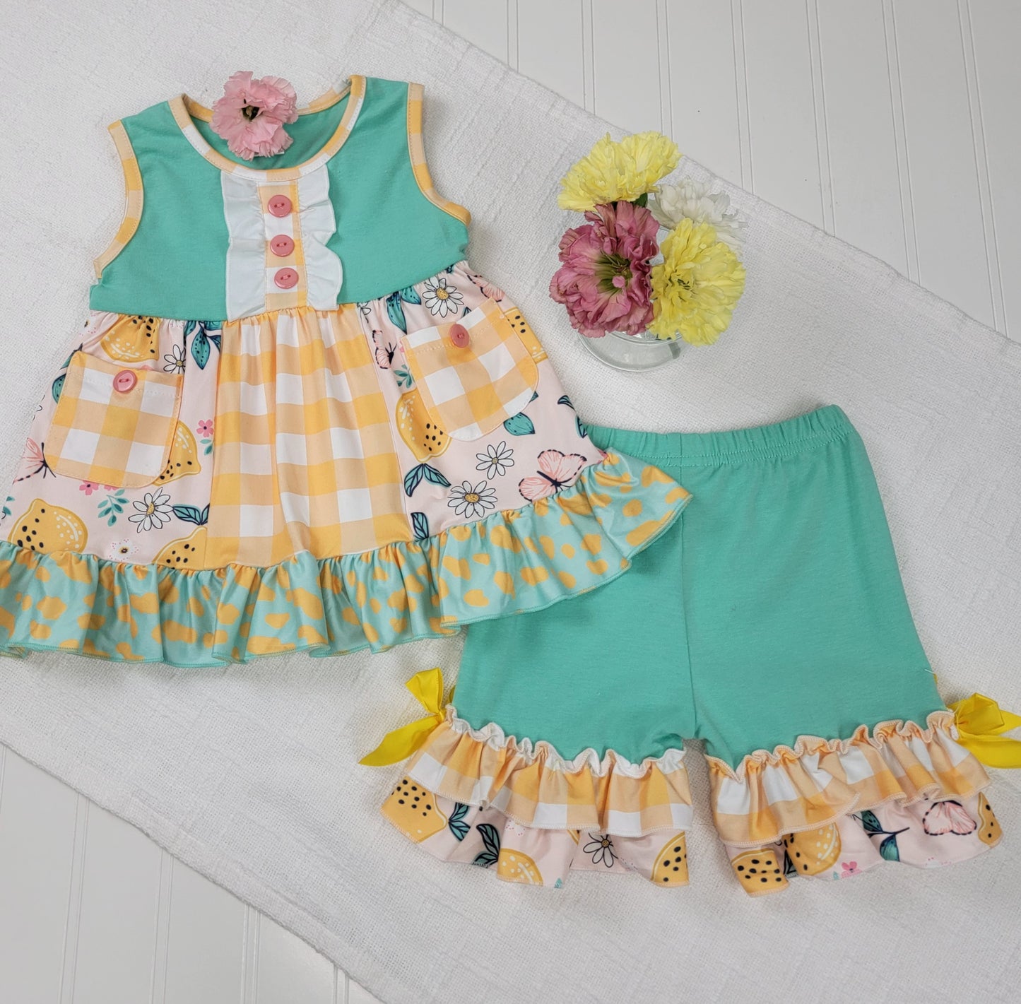 The Melina & Me Baby: Citrus Outfit is a toddler girl’s two-piece set with a sleeveless teal and yellow gingham lemon print dress with ruffles, plus matching ruffle-hem teal shorts, displayed on a white textured surface.