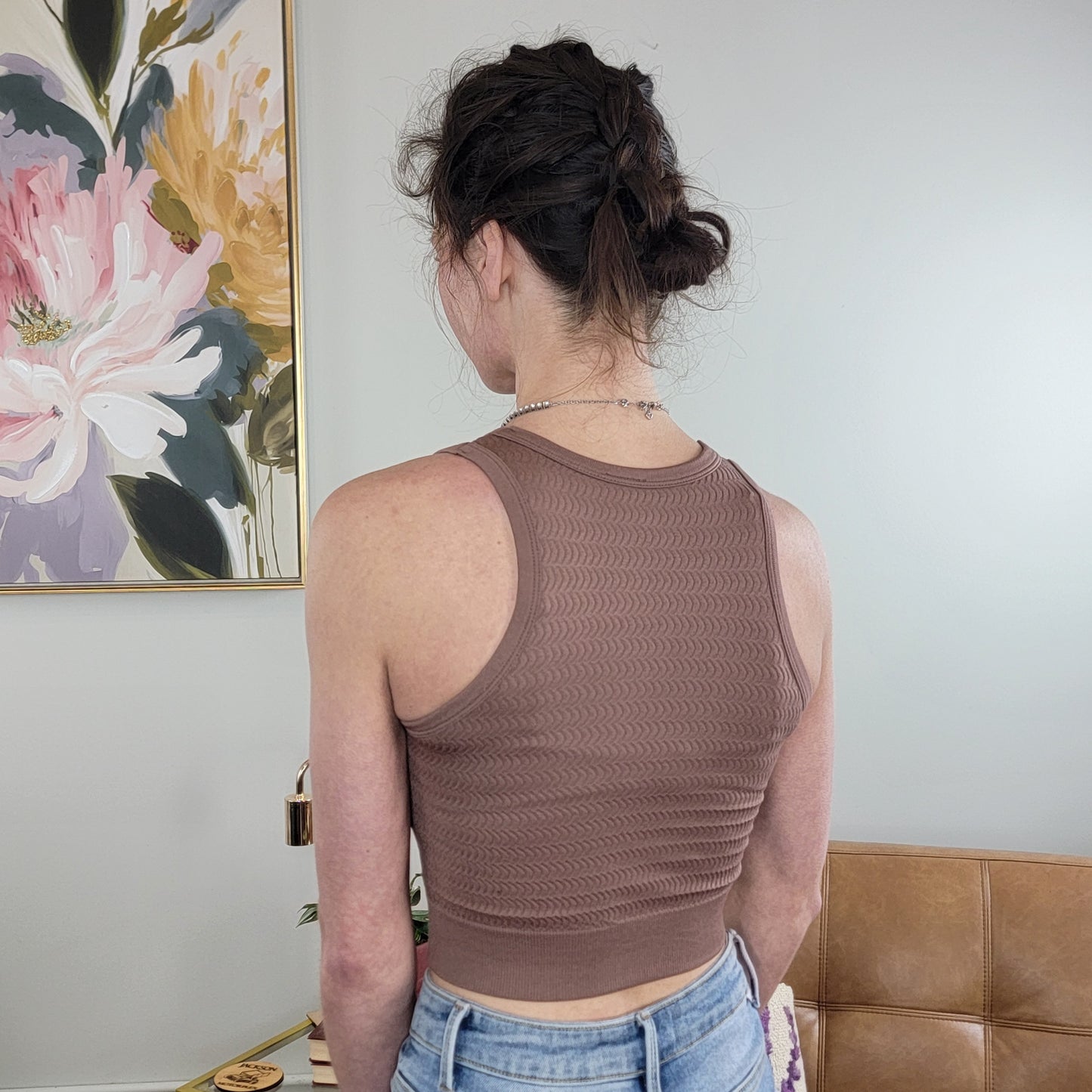 A woman with dark hair in a braided updo models the Superline Suzanne Textured Tank in mocha and light blue jeans, standing with her back to the camera near a floral painting and a tan couch.