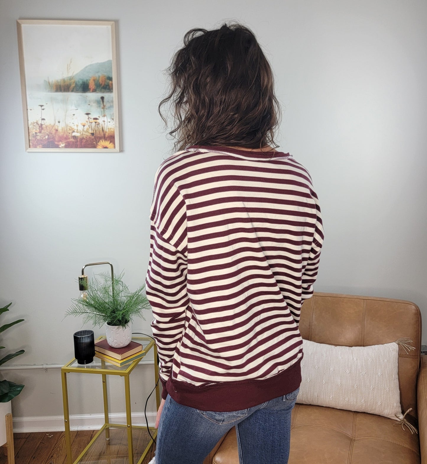 A person with wavy brown hair, wearing the Staccato Lucia Striped Pullover and blue jeans, stands facing away in a cozy room featuring a tan chair, a small table with a plant, and a picture on the wall.