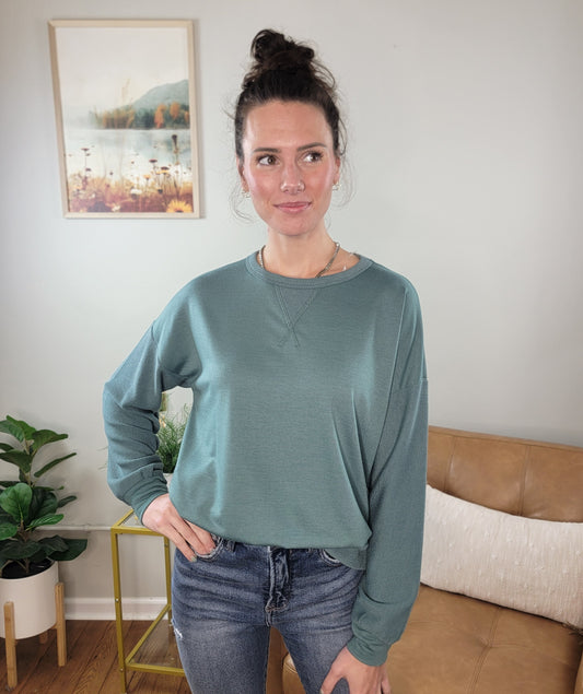 A woman with dark hair in a bun poses indoors, wearing the Staccato Paityn Long Sleeve Top in sage green and blue jeans. She stands by a tan couch, with plants and a landscape painting behind her.