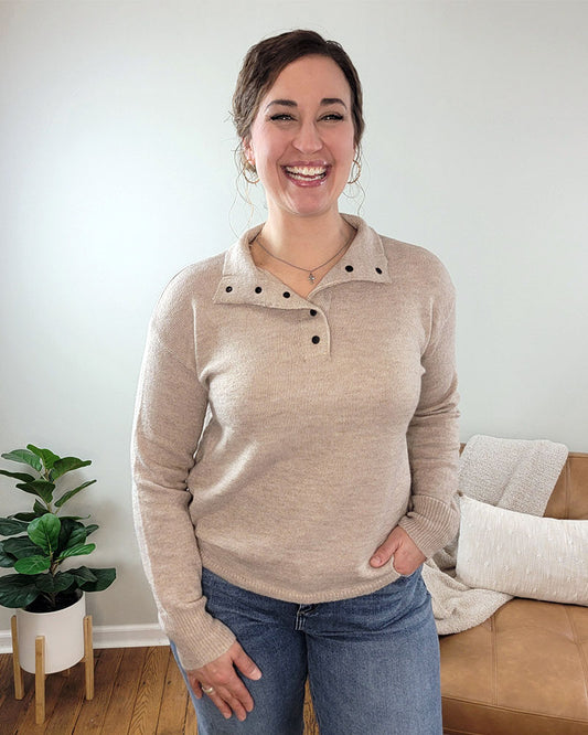 A woman with brown hair smiles indoors, wearing the Staccato Devon Half Snap Sweater in taupe and blue jeans, one hand in her pocket. Behind her is a tan sofa with a light blanket and a potted plant on the wooden floor.