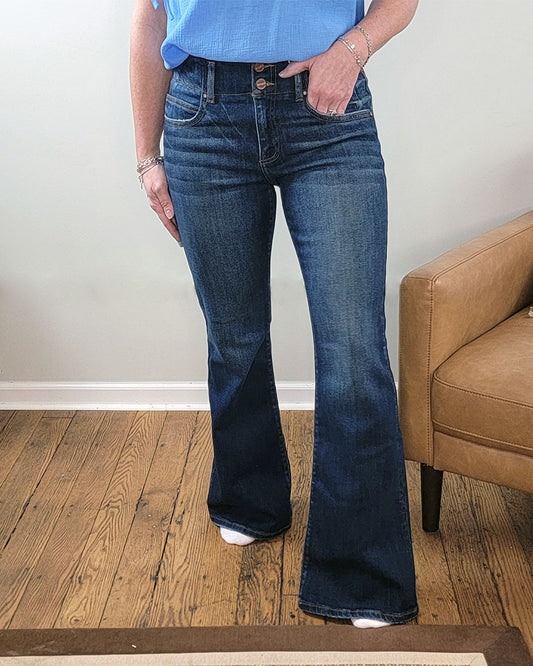 A person wears Risen's Eleanor Flare Jeans indoors, standing on a wooden floor near a tan armchair, with only their lower half visible and hands resting by their waist.