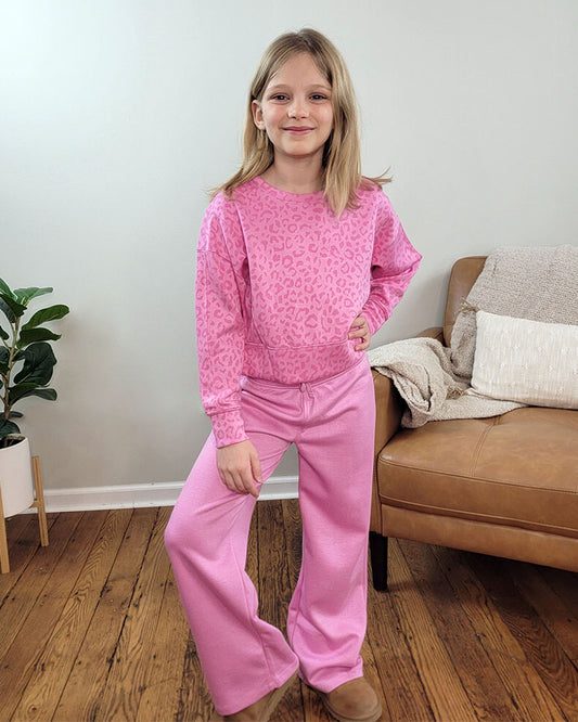 A young girl stands indoors on a wooden floor, smiling with one hand on her hip. She wears the Reflex Girls Jessi Leopard Set, featuring a leopard-print pullover and wide-leg pants. Behind her is a tan couch with cream blankets and a green potted plant.