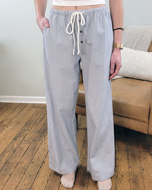 A person stands barefoot on a wooden floor, wearing Love Poem's Vedea Pinstripe Pant in grey-blue with a white top. A tan couch with cushions is visible in the background.