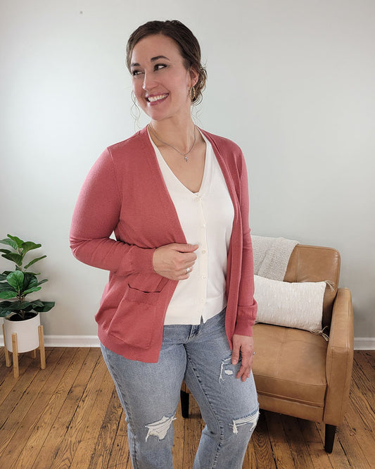 A woman smiles in a cozy room, wearing Staccato's Luna Button Down Cardigan in cream and rose over a white blouse and ripped jeans. Behind her are a tan armchair with a pillow and a potted plant on the hardwood floor.