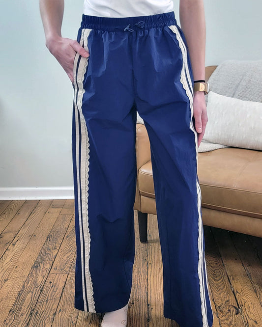A person stands indoors on a wooden floor, wearing Veveret's Scarlett Striped Lace Pants featuring a navy color, elastic waistband, white side stripes, and scalloped trim; a beige couch with light pillows appears in the background.