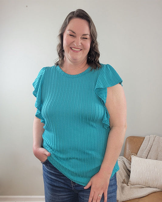 A woman with wavy brown hair smiles indoors against a light wall, wearing the Heimish Havanah Ruffle Sleeve Top in turquoise with blue jeans. A beige couch topped with a blanket is seen in the background.
