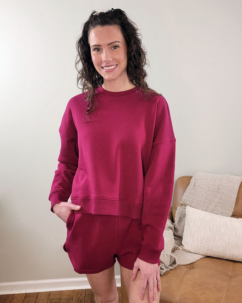 A woman with curly hair smiles while wearing the ZENANA Aria Sweatshirt + Shorts Set, standing indoors by a tan couch with pillows and a knitted blanket.