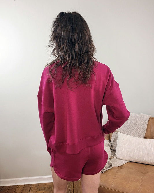 A person with wavy brown hair stands indoors facing away, wearing the magenta Aria Sweatshirt + Shorts Set by ZENANA, near a tan couch with light pillows and a blanket.