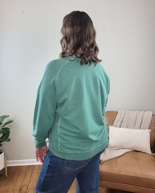 A person with wavy brown hair, wearing the ZENANA Nelly Pullover Crewneck in dark green and blue jeans, stands with their back to the camera in a living room featuring a tan couch, blanket, cushion, and a potted plant.