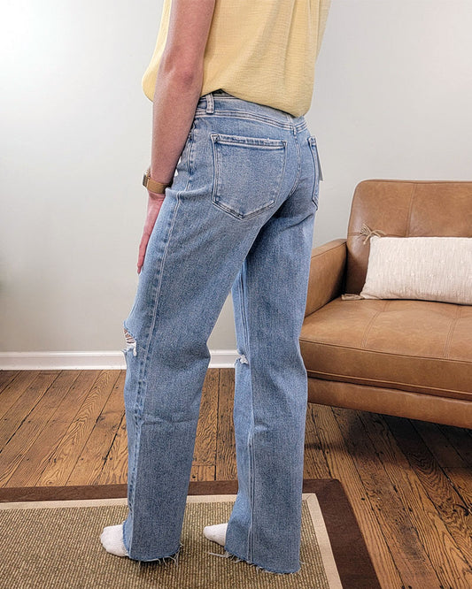A person stands indoors on a rug, wearing Glorietta Slim Wide Leg Jeans from Lovervet, a yellow short-sleeve shirt, and white socks. In the background is a tan leather couch with a white pillow.