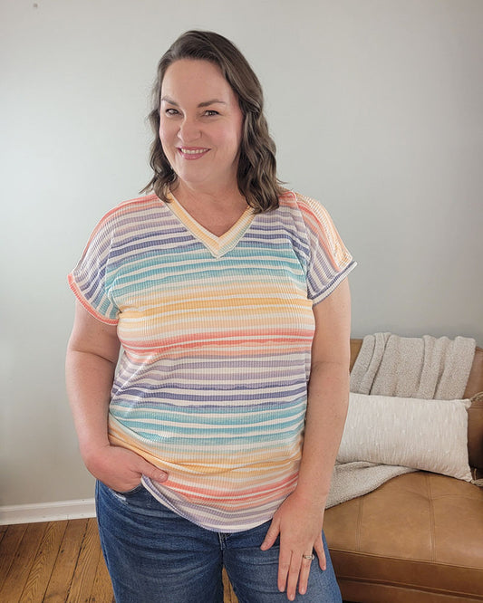 A woman with wavy brown hair smiles at the camera indoors, wearing the Heimish Hilary Striped V-Neck and blue jeans. One hand is in her pocket, and a tan couch with a blanket appears in the background.