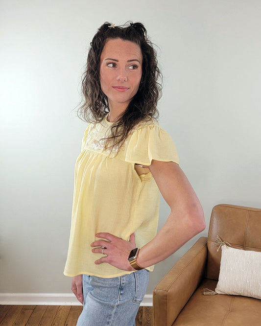 A woman with curly brown hair wears the Mine and E&M Addison Flutter Sleeve Top in butter yellow and blue jeans, standing indoors by a tan armchair, looking left with one hand on her hip.