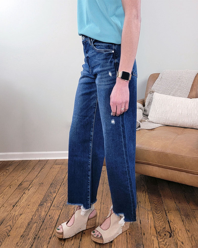 Indoors on a wooden floor, a person wears a light blue top, Risen’s Briar Crop Wide Leg Jeans in dark wash with frayed hems, beige wedge sandals, and a smartwatch. A tan couch with pillows and a blanket is in the background.