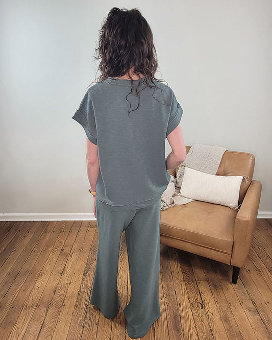 A person with wavy brown hair, facing away, wears the ZENANA Daphne Top and Pant Set—a lightweight cotton loungewear outfit—on a wooden floor near a tan armchair with a white/beige pillow and throw. Perfect for cozy travel.