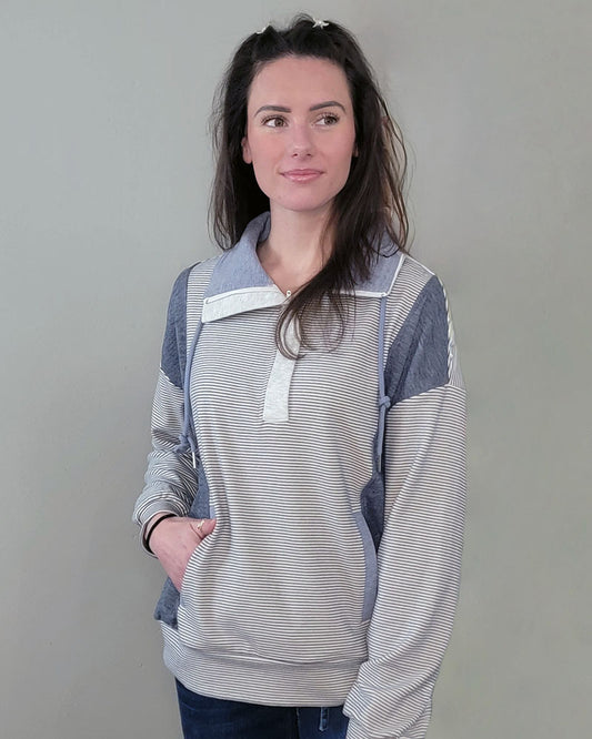 A woman with long brown hair wears the Hem & Thread Shay Striped Mix Pullover in light gray and white, standing against a plain gray wall. She looks slightly right with a neutral expression, one hand casually in her pocket.