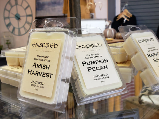 Close-up of two Enspired Soy Wax Melts, labeled Amish Harvest and Pumpkin Pecan, displayed among other wax melts and decor in a shop with a bar deals sign in the background.