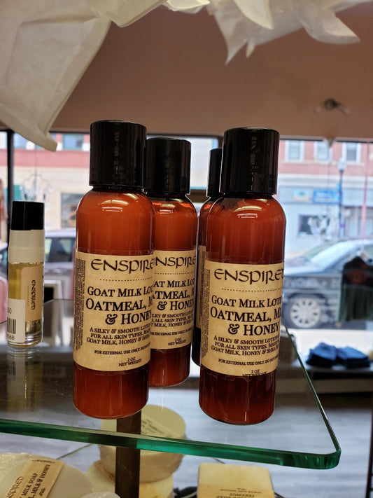 Three bottles of Enspired Hand Lotion labeled “Oatmeal, Milk & Honey” are displayed on a glass shelf in a store with a city street visible through the window behind them.