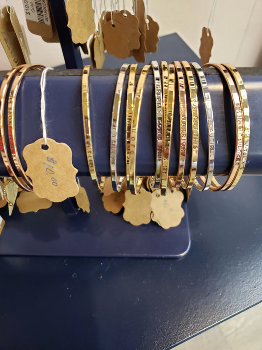 LA Jewelry House Inspiration Bracelets in gold and silver with engraved text are displayed on a dark stand. Each bracelet has a brown price tag, with $12.00 visible on one of the tags.