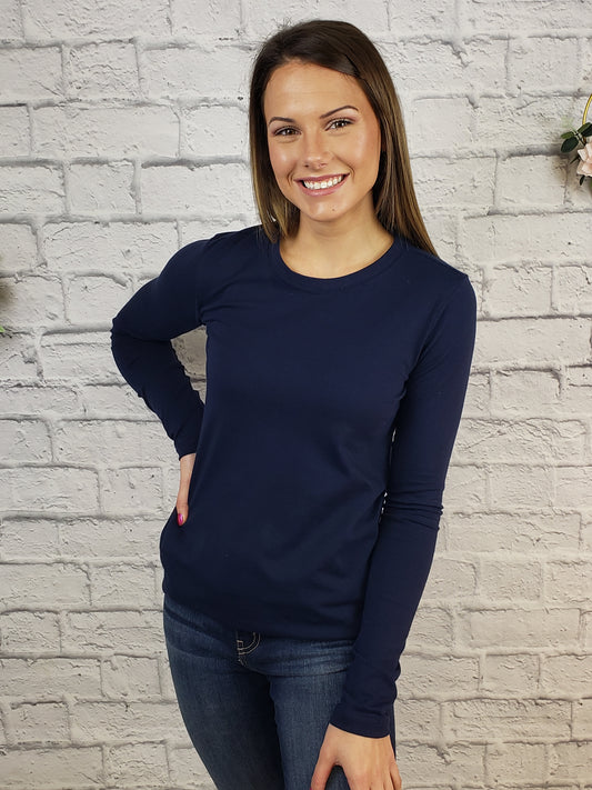 A woman with long brown hair smiles in front of a white brick wall, wearing the Zenana Basic Long Sleeve Crew in navy blue and blue jeans, with her right hand on her hip.