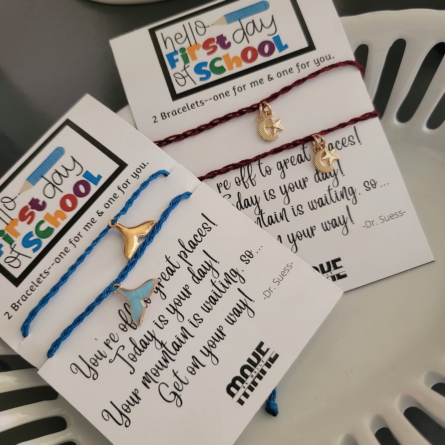 Two "Make By Molly: Charm Bracelet Sets" by Molly Thurmer, each on a card with "Hello, first day of school" and a Dr. Seuss quote. Includes one blue whale tail and one red crescent moon bracelet, in a white tray.