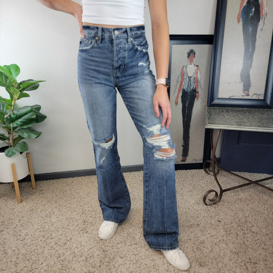 A person wearing a white sleeveless top, KanCan's Indya Vintage 90's Flare jeans, and white sneakers stands on a beige carpet with a green plant and framed fashion artwork in the background.
