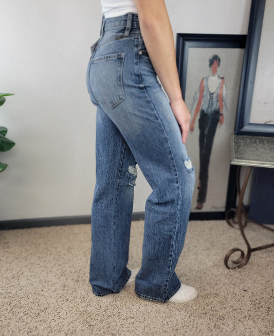 A person in high-waisted blue KanCan Indya Vintage 90's Flare jeans and white sneakers stands on a beige carpet beside a framed fashion illustration of a woman dressed similarly.