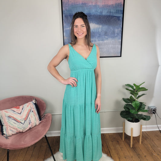 A smiling woman with long brown hair wears the Timing Vivian Strappy Maxi Dress and stands indoors by a pink chair with a pillow, a potted plant, and abstract wall art in the background.
