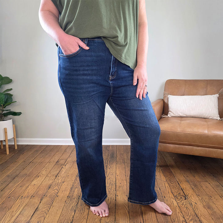 A person wearing a green top and Katelyn crop jeans by Risen stands barefoot on a wooden floor in a living room with a tan sofa, pillow, and plant in the background.