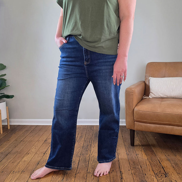 A person stands barefoot on a wooden floor, wearing Risen's Katelyn crop jeans in dark blue and an olive green t-shirt, with their left hand in their pocket. A light brown armchair and a plant are visible in the background.