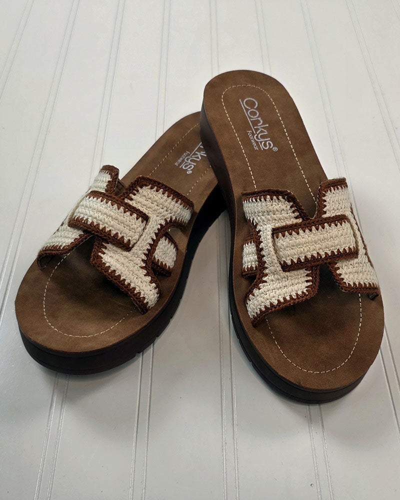 A pair of brown Corkys On the Edge Sandals with white and brown geometric woven straps, displayed on a white wooden surface. Stylish and versatile, these summer slide sandals are a must-have for any wardrobe.
