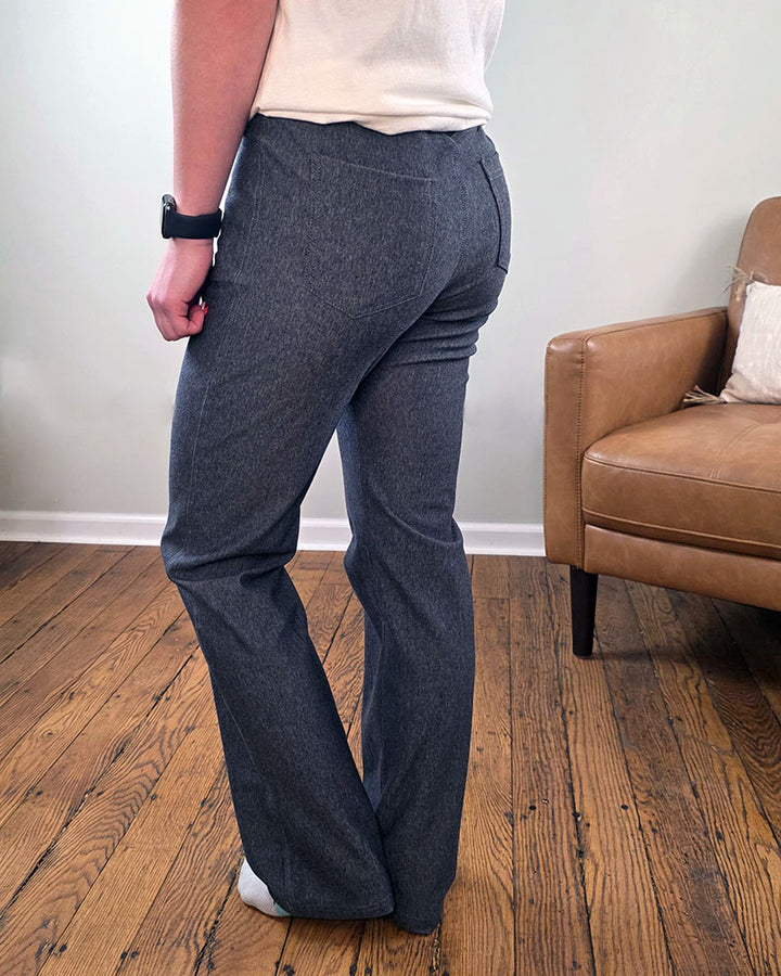 A person stands indoors on a wooden floor wearing stylish gray Blaire Straight Leg Jeggings by Yelete Group, a white top, black wristwatch, and white socks. Behind them is a tan armchair with a white pillow against a light-colored wall.