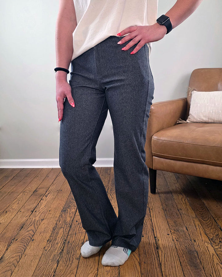 A person wearing Blaire Straight Leg Jeggings by Yelete Group and a white shirt stands on a wooden floor next to a tan chair. Their nails are painted pink, and they wear light-colored socks for a modern, stylish look.