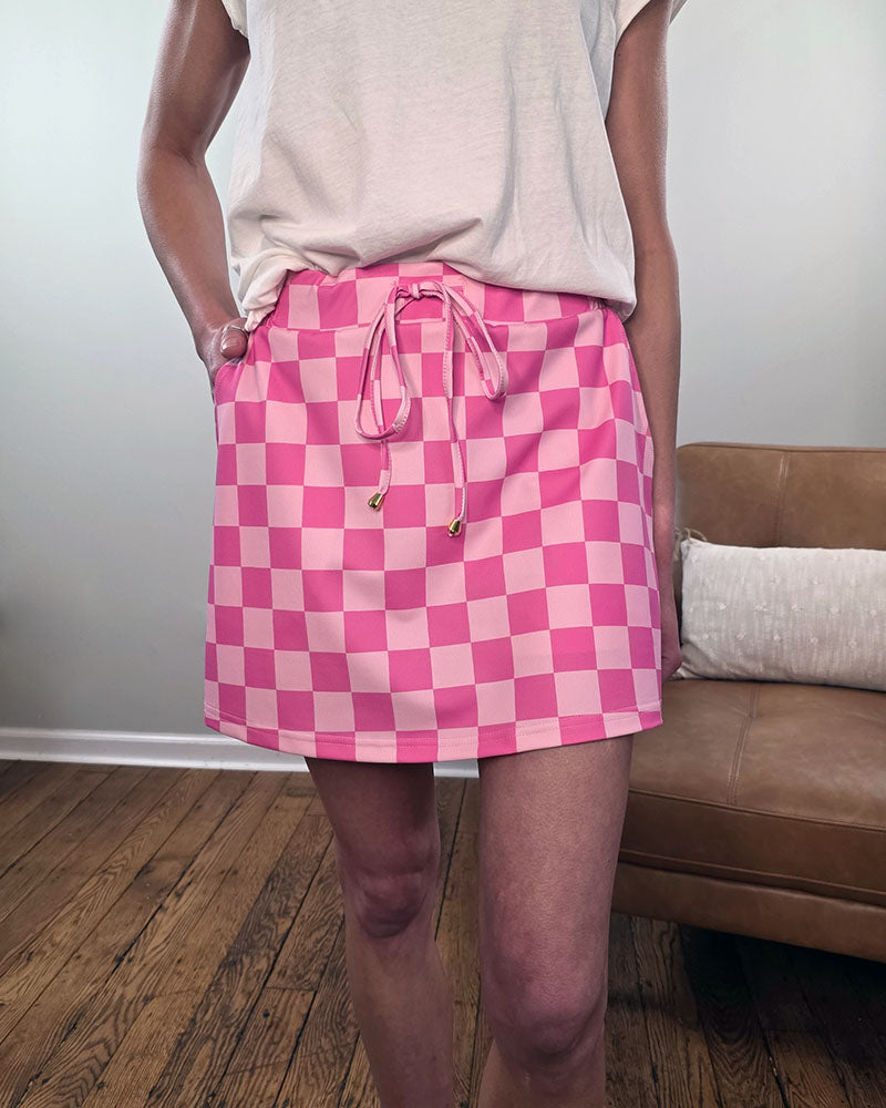 A person stands indoors wearing a white shirt and Jess Lea's Everyday Skorts - Stylish Patterns for Summer in a pink-and-white checkered design. In the background are a brown couch, a white pillow, and a wooden floor.
