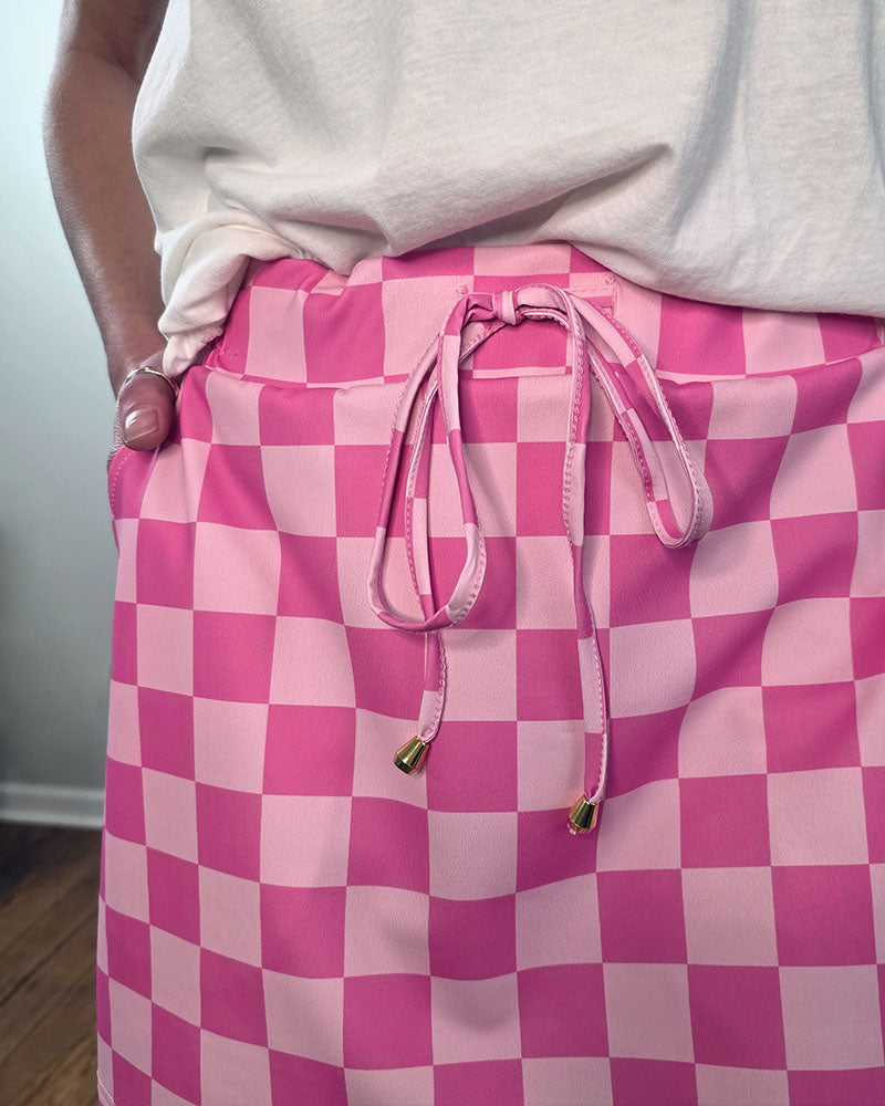A person wearing a white top and the Jess Lea Everyday Skort—a pink checkerboard skirt with a drawstring waist and gold metal tips—stands confidently with one hand in their pocket, embodying easy summer style.