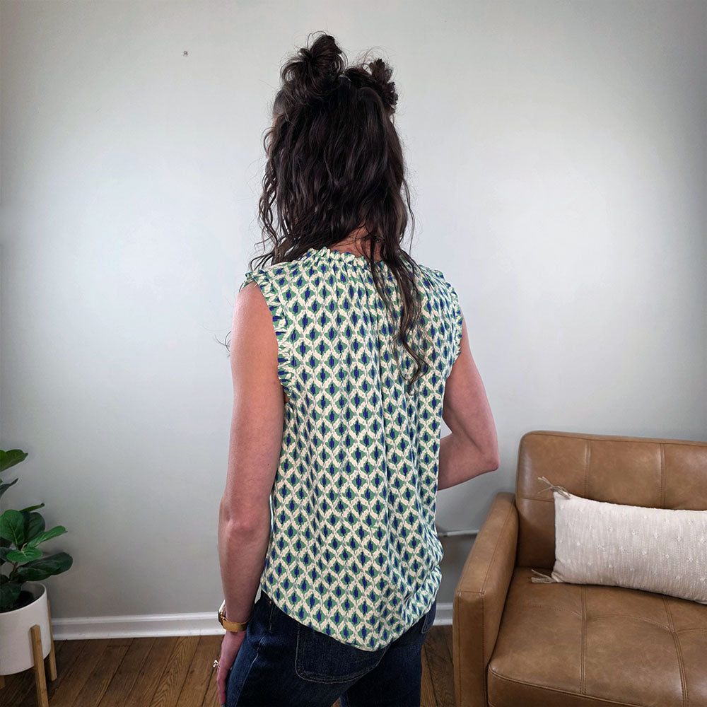 A person with long, wavy brown hair stands indoors wearing the Staccato Marcella Sleeveless Patterned Top for Work and dark jeans. A tan couch, pillow, and potted plant create a stylish setting—perfect work wardrobe inspiration.