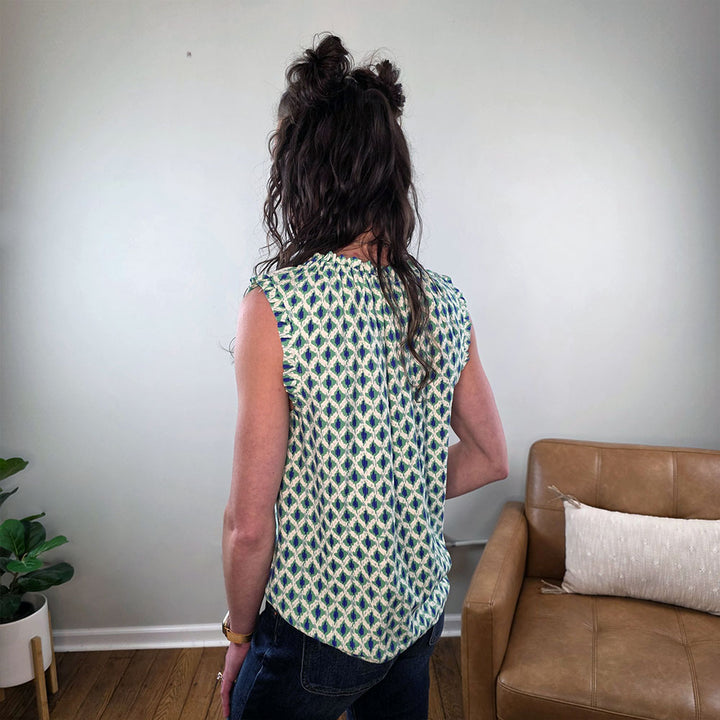 A person with long, wavy brown hair stands indoors wearing the Staccato Marcella Sleeveless Patterned Top for Work and dark jeans. A tan couch, pillow, and potted plant create a stylish setting—perfect work wardrobe inspiration.