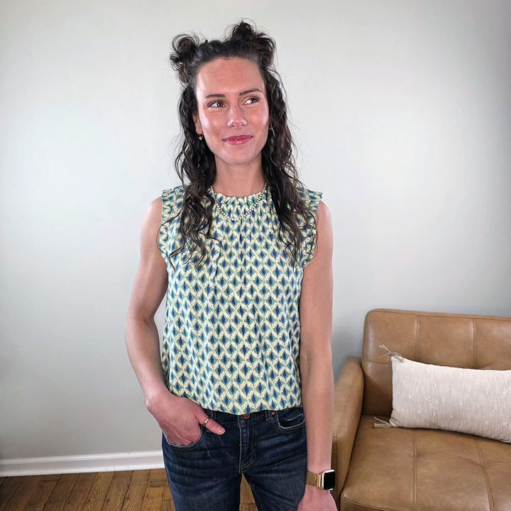 A woman with long curly hair in two buns smiles indoors, wearing the Staccato Marcella Sleeveless Patterned Top for Work and jeans. She stands next to a tan couch—a chic choice to enhance your work wardrobe.