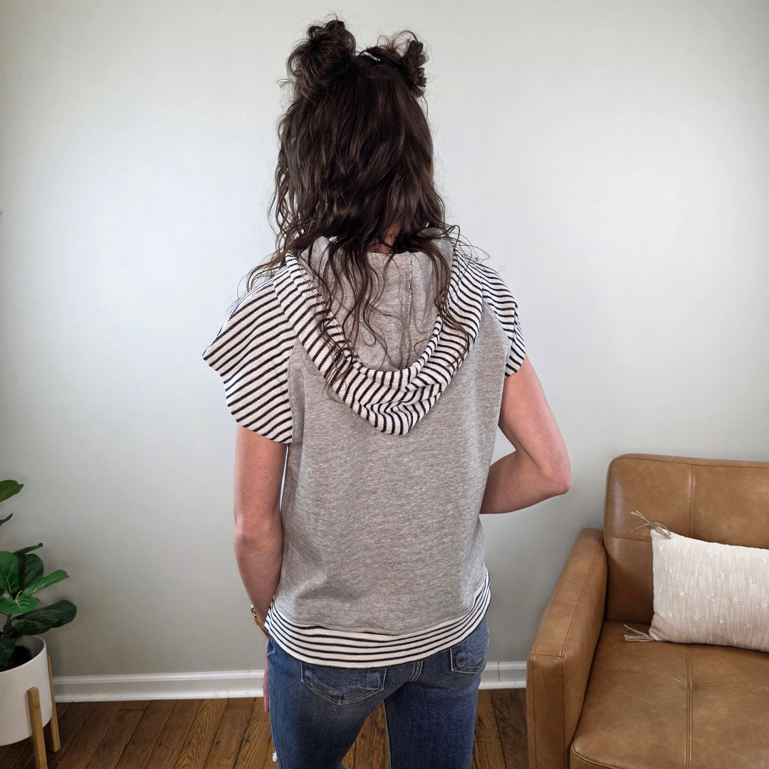 A person with wavy brown hair in two buns stands with their back to the camera, wearing the Staccato Genise Short Sleeve Hooded Casual Lounge Top in gray, blue jeans, in a cozy room with a tan couch and a plant.