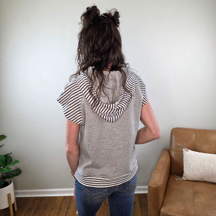 A person with wavy brown hair in two buns stands with their back to the camera, wearing the Staccato Genise Short Sleeve Hooded Casual Lounge Top in gray, blue jeans, in a cozy room with a tan couch and a plant.