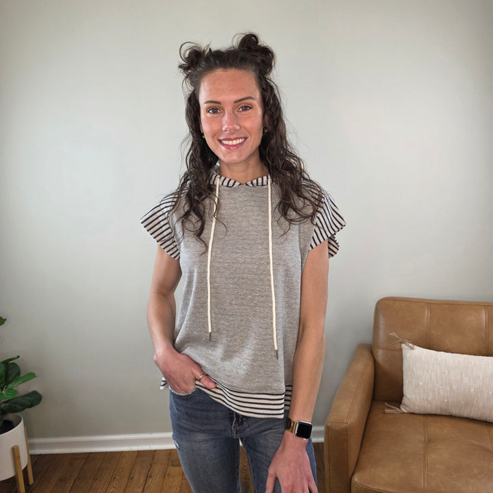 A woman with wavy brown hair styled in half-up buns smiles at the camera, wearing the Staccato Genise Short Sleeve Hooded Casual Lounge Top with jeans and an Apple Watch, embodying relaxed style in a cozy, neutral-toned room.