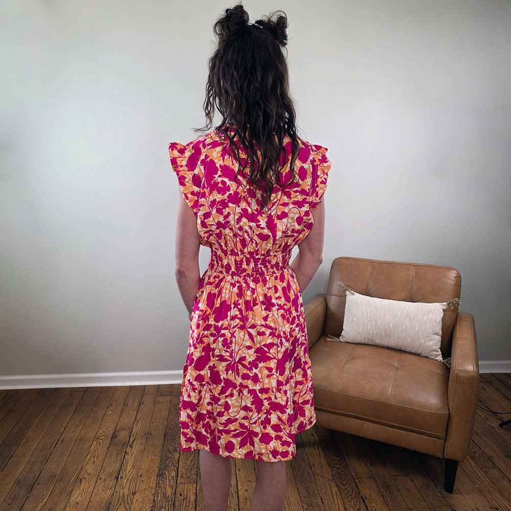 A person with wavy dark hair in two buns, seen from behind, wears the Staccato Edith Dress—a bright floral piece with ruffle details—in a room featuring wooden floors, a tan armchair, and a light-colored pillow.