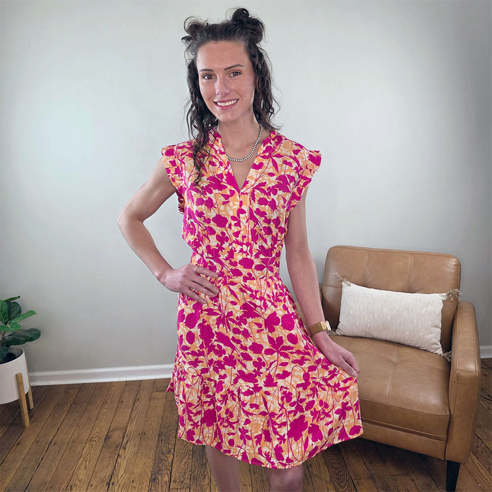A woman stands indoors on wooden flooring, smiling at the camera in the bright floral Edith Dress by Staccato. One hand rests on her hip. A tan armchair with a pillow and a green potted plant are visible in the background.