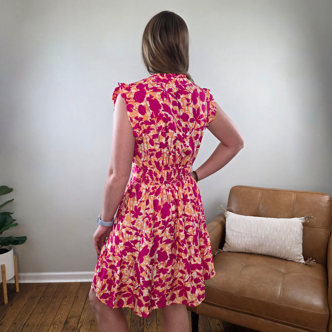 A woman stands indoors with her back to the camera, wearing the Staccato Edith Floral Print Sleeveless Dress. She poses beside a tan leather chair and pillow in a room with wooden floors and light-colored walls.