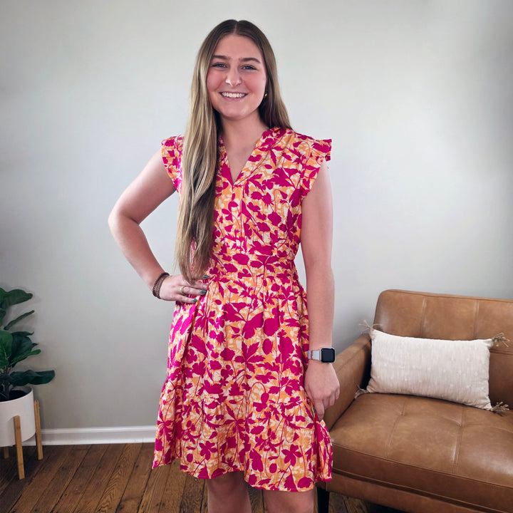 A young woman with long brown hair wears the Staccato Edith Floral Print Sleeveless Dress and smiles in a living room featuring wooden floors, a tan chair, and a potted plant in the background.