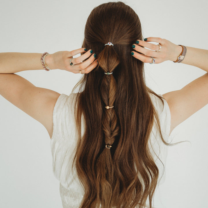 A woman with long brown hair styled in a bubble braid holds her hair with both hands, showcasing her collection of K’lani hair tie bracelets. She wears the “Terra” Earth Tones Hair Tie Rings as both stretchy rings and bracelets, effortlessly pairing these versatile accessories with her sleeveless white top against a plain white background.