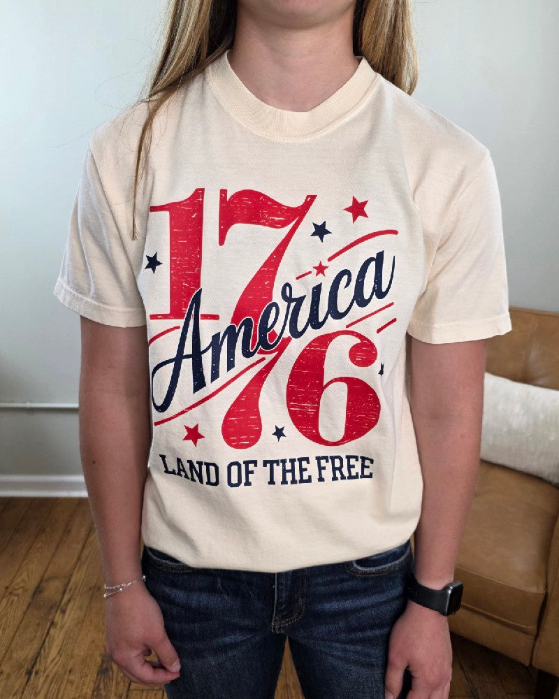 A person wearing the cream Land of the Free 1776 Graphic Tee by Cosmos + Clover Boutique, featuring bold red and blue text, stands in a room with a wooden floor, brown couch, and white pillow.